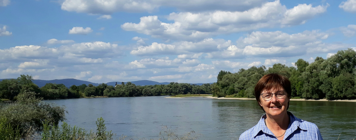 Das Bild zeigt die Donau mit den Donauauen bei blauem Himmel mit einigen Wolken. Rechts im Bild lächelt MdL Rosi Steinberger in die Kamera.