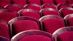 Das Bild zeigt fünf Stuhlreihen eines Theateres in Großaufnahme. Die Stühle und Polster sind in Weinrot gehalten.